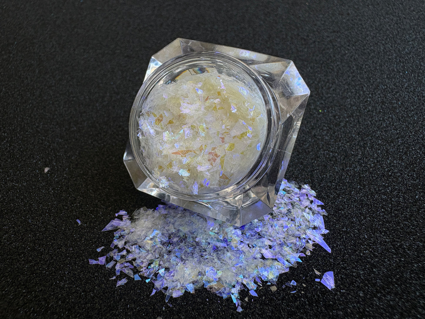 Iridescent white cellophane glitter flakes with purple reflections spilled from a clear jar onto a black textured background.