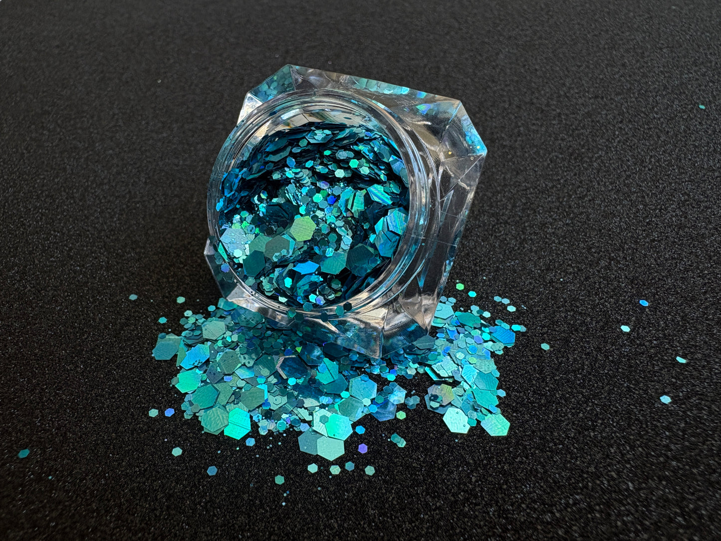 Super chunky holographic dark aqua glitter mix in a clear jar, spilled onto a black textured background.