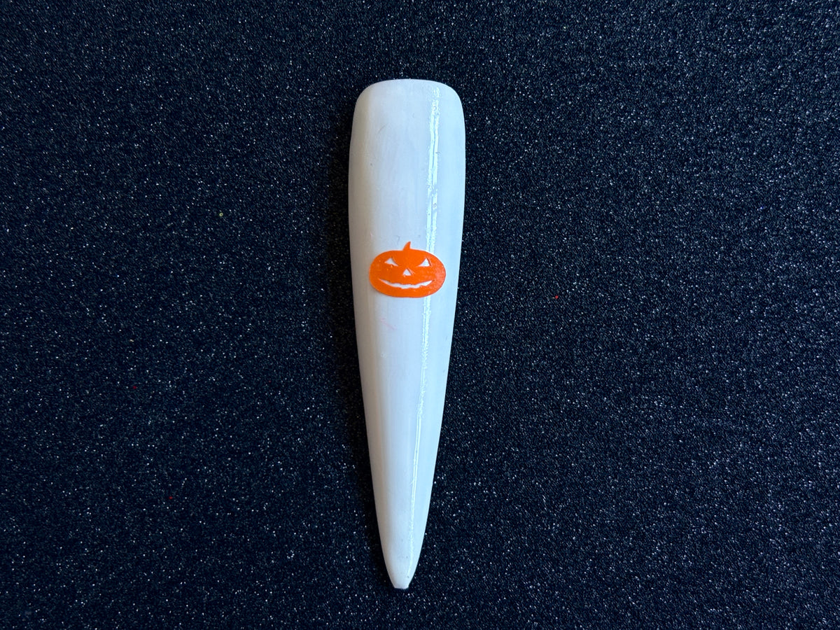 Orange pumpkin Halloween nail sticker applied on a glossy white stiletto nail tip against a black textured background.