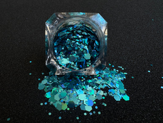 Super chunky holographic dark aqua glitter mix in a clear jar, spilled onto a black textured background.