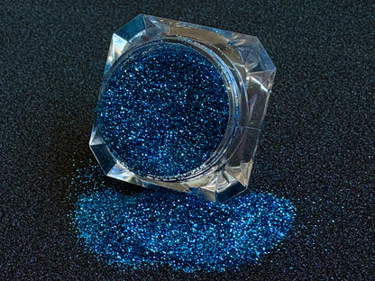 Blue and teal glitter mix spilled from a clear jar onto a black textured background.