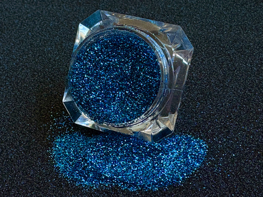 Blue and teal glitter mix spilled from a clear jar onto a black textured background.