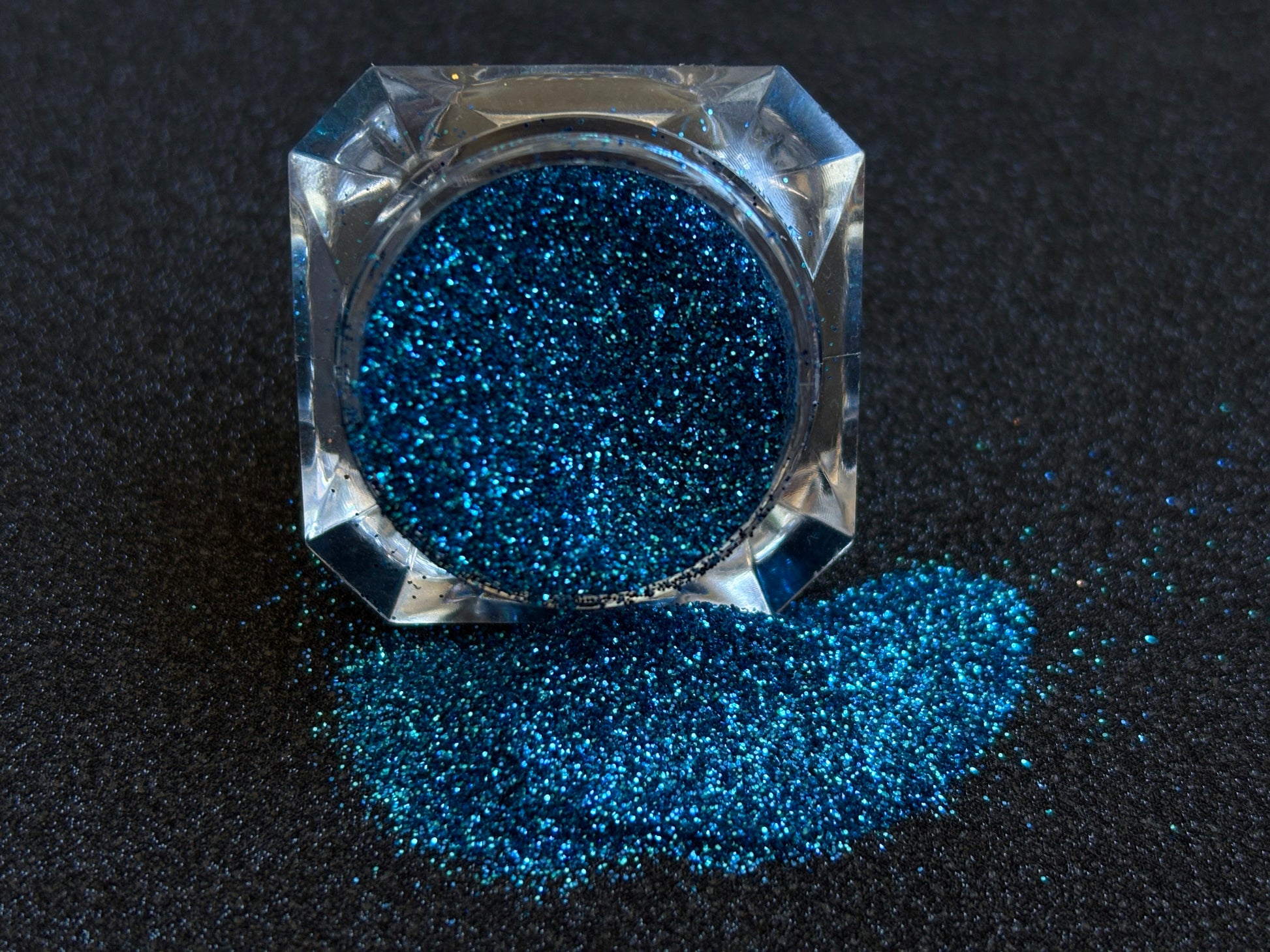 Blue and teal glitter mix spilled from a clear jar onto a black textured background.