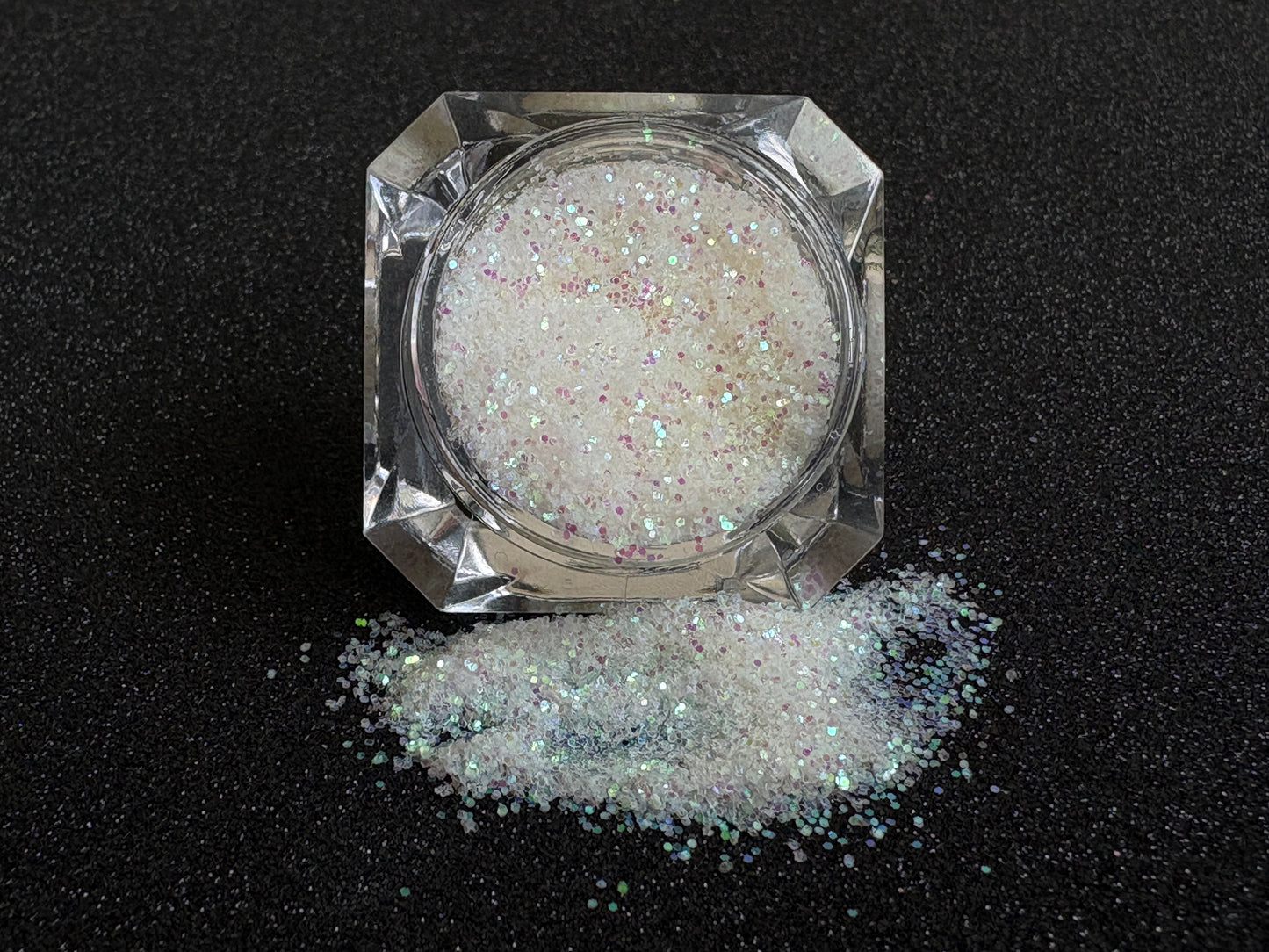 Iridescent white glitter spilled from a clear jar onto a black textured background.