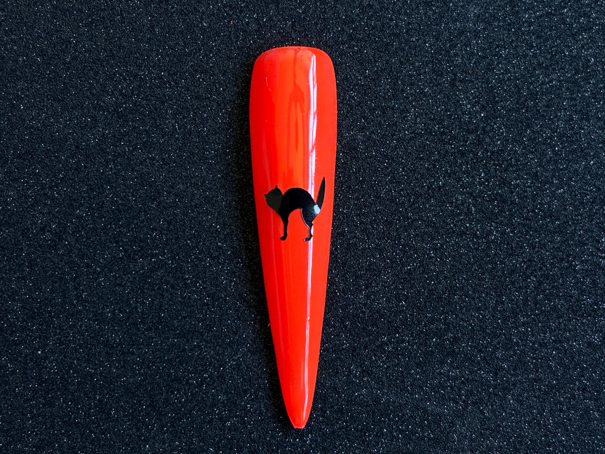 Black cat Halloween nail sticker applied on a bright orange stiletto nail tip against a black textured background.