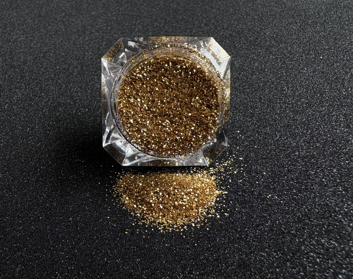 Gold metallic glitter spilled from a clear jar onto a black textured background.