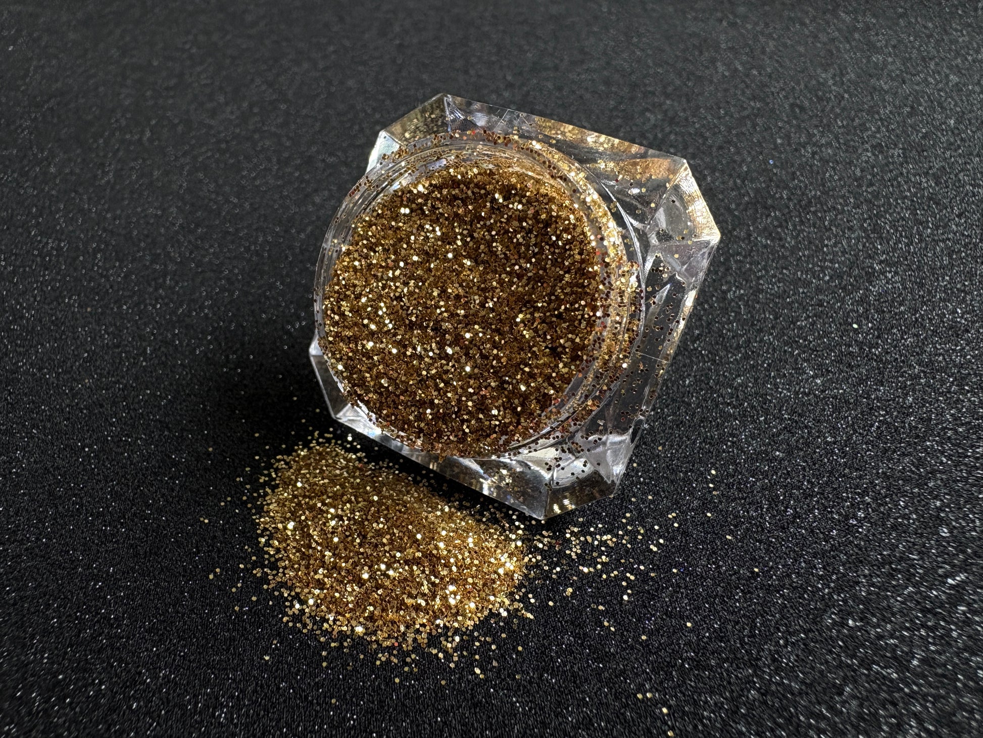 Gold metallic glitter spilled from a clear jar onto a black textured background.