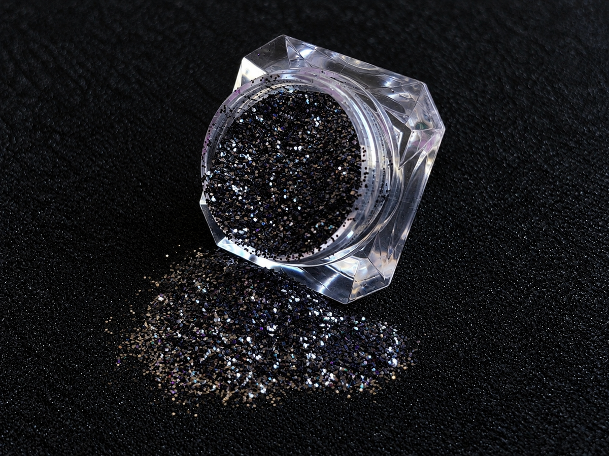 Holographic black glitter spilled from a clear jar onto a black textured background.