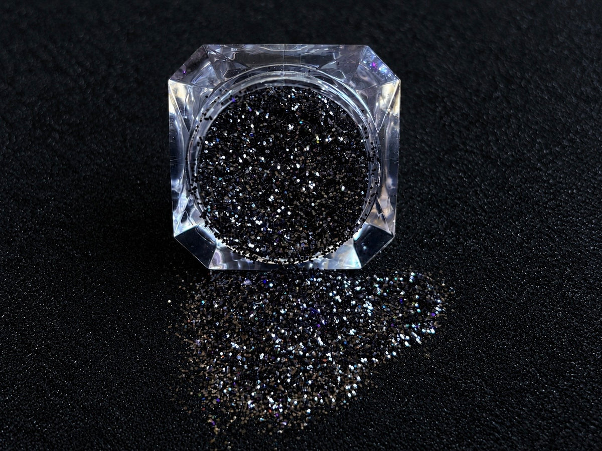 Holographic black glitter spilled from a clear jar onto a black textured background.