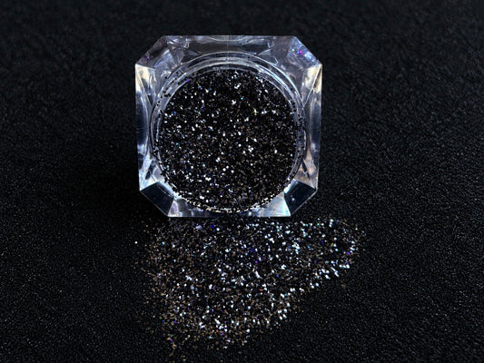 Holographic black glitter spilled from a clear jar onto a black textured background.