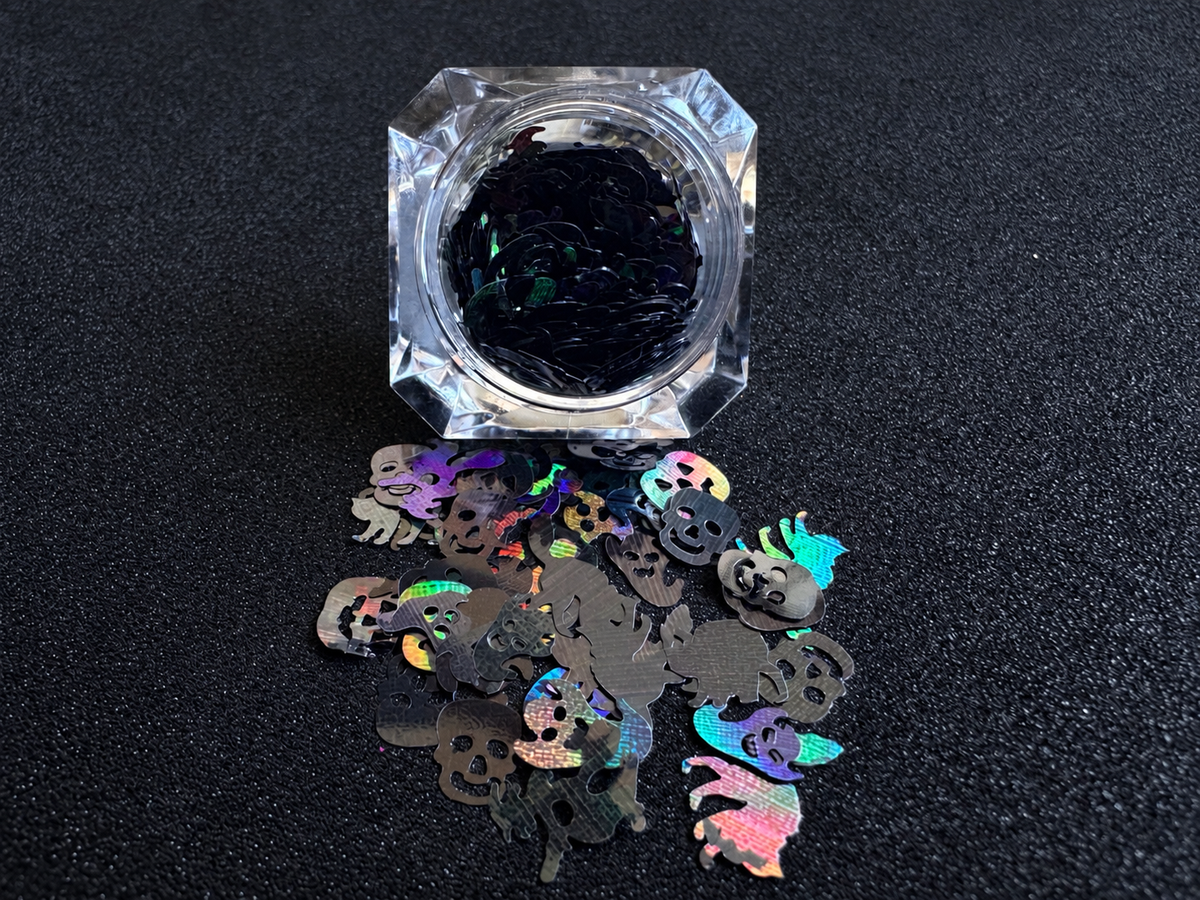 Holographic black Halloween glitter shapes featuring pumpkins, skulls, ghosts, cats, bats, and witches spilled from a clear jar onto a black textured background.
