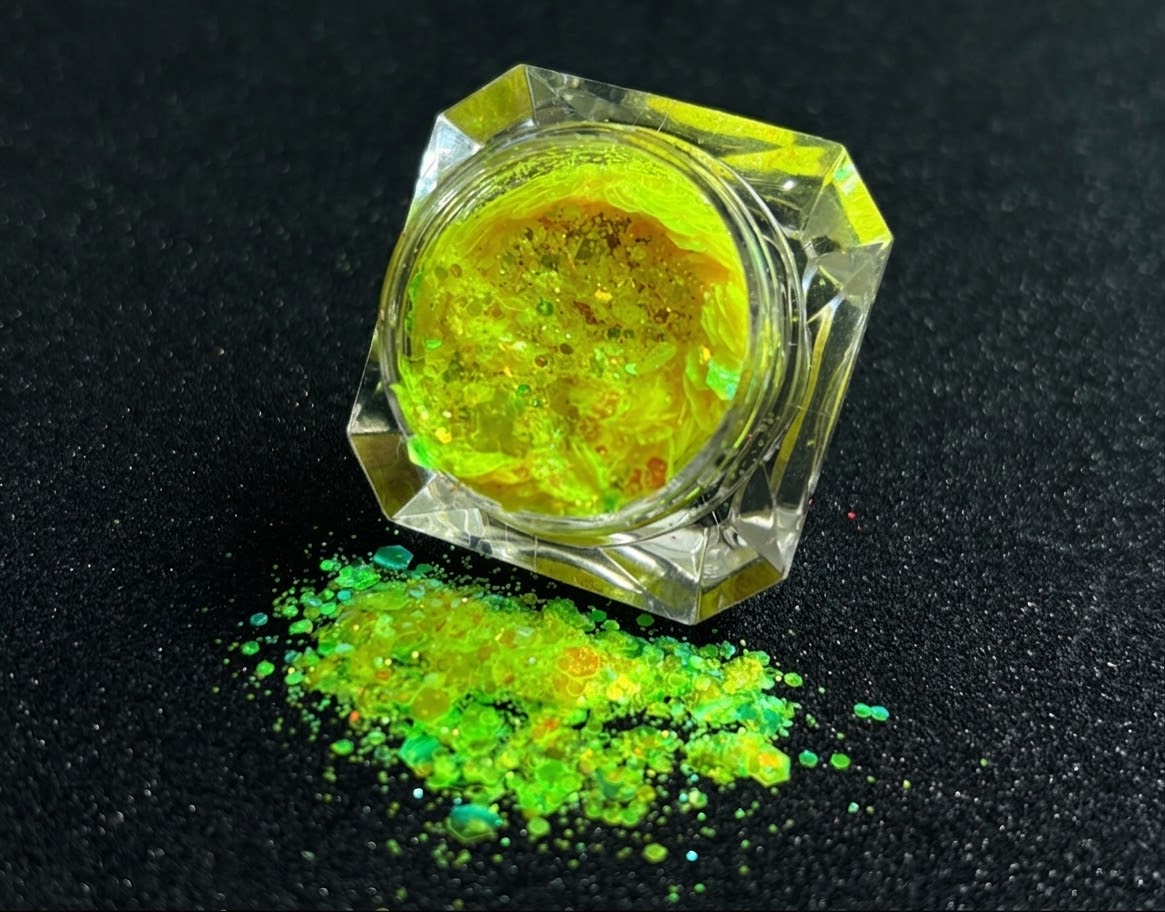 Fluorescent iridescent green super chunky glitter mix with yellow shift spilled from a clear jar onto a black textured background.