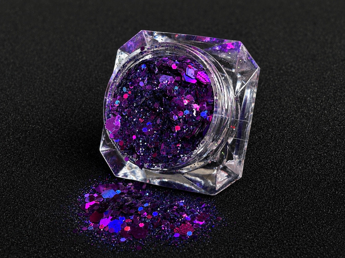 Deep purple and violet super chunky glitter mix with holographic sparkle spilled from a clear jar onto a black textured background.