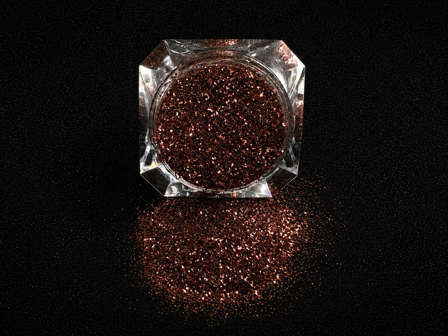 Chocolate brown metallic glitter spilled from a clear jar onto a black textured background.