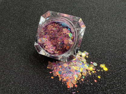 Top-down view of super chunky pastel purple glitter mix with rose gold iridescent color shift, spilled from a clear jar onto a black textured background.