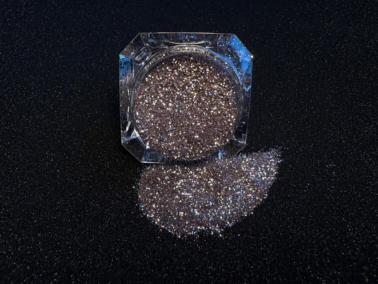 Champagne pink glitter spilled from a clear jar onto a black textured background.