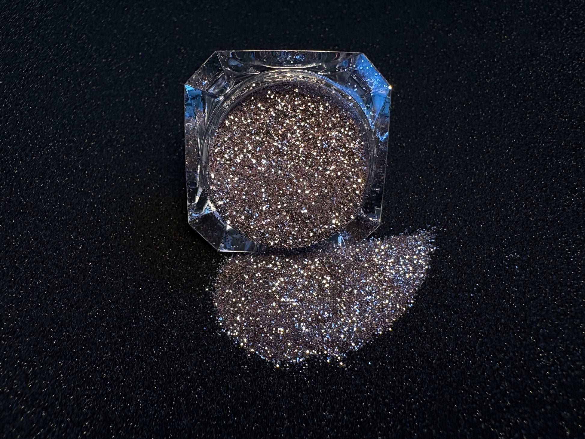 Champagne pink glitter spilled from a clear jar onto a black textured background.