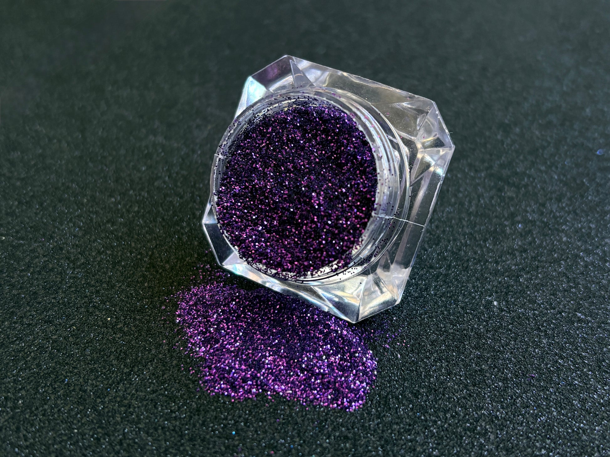 Royal purple metallic glitter spilled from a clear jar onto a black textured background.