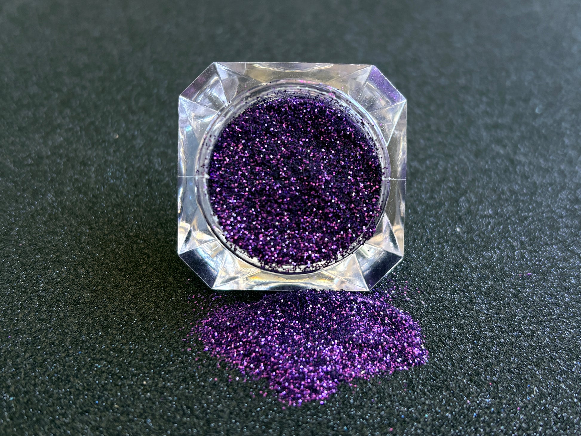 Royal purple metallic glitter spilled from a clear jar onto a black textured background.