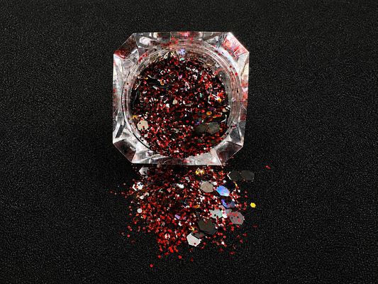 Ruby red, black, silver, and gold holographic super chunky glitter mix spilled from a clear jar onto a black textured background. 