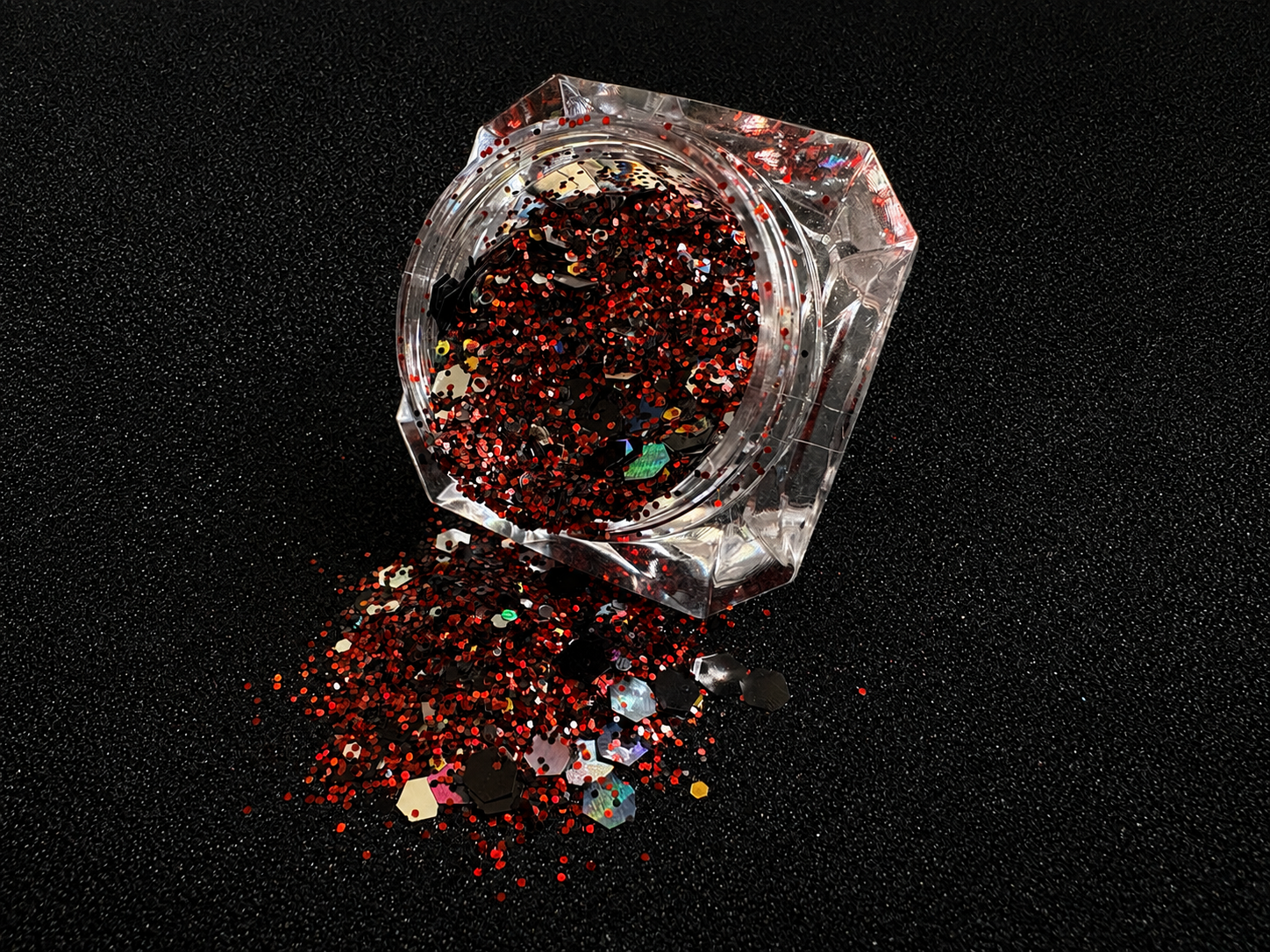 Ruby red, black, silver, and gold holographic super chunky glitter mix spilled from a clear jar onto a black textured background. 