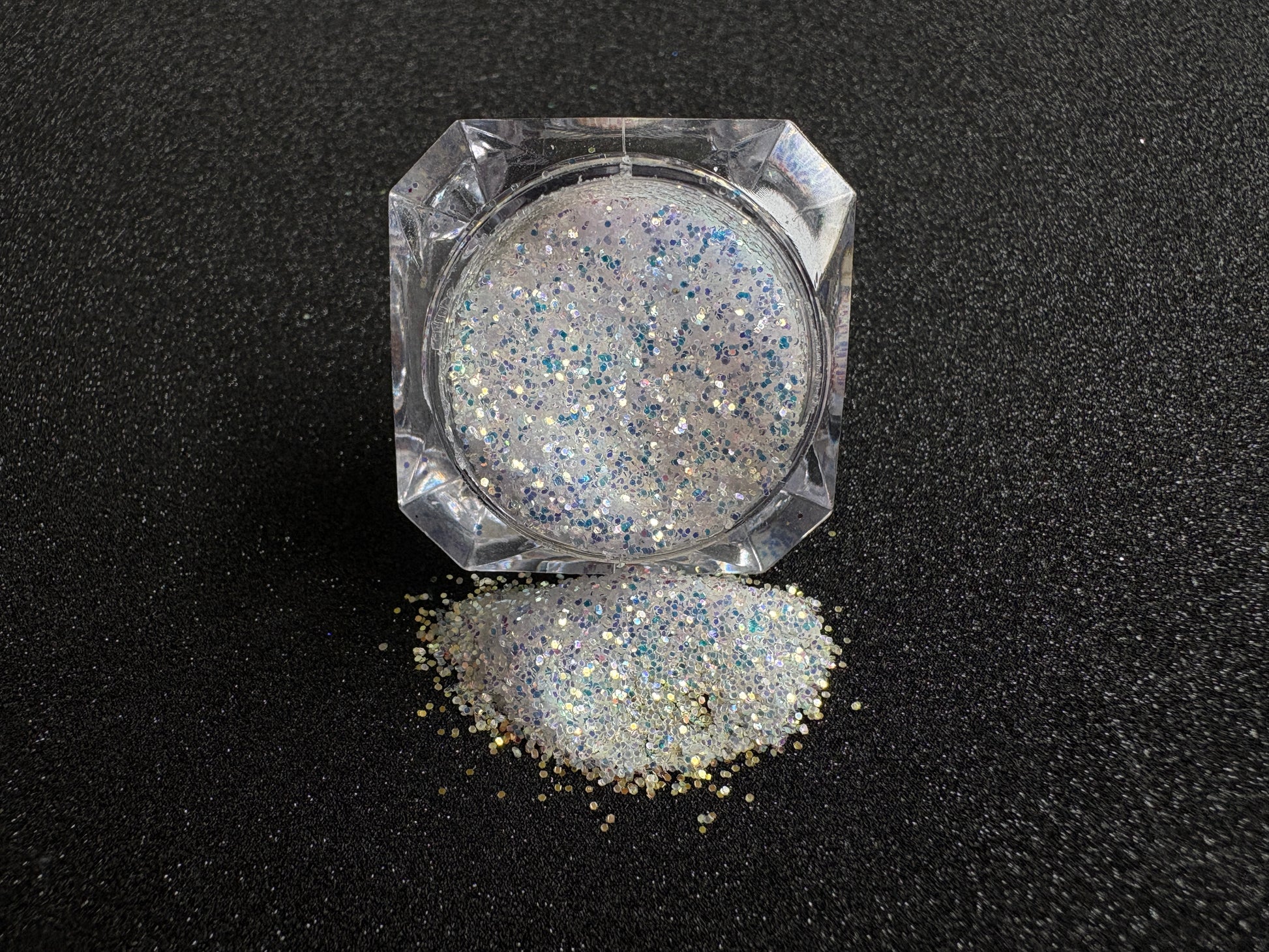 Blue-purple and gold glitter mix spilled from a clear jar onto a black textured background.
