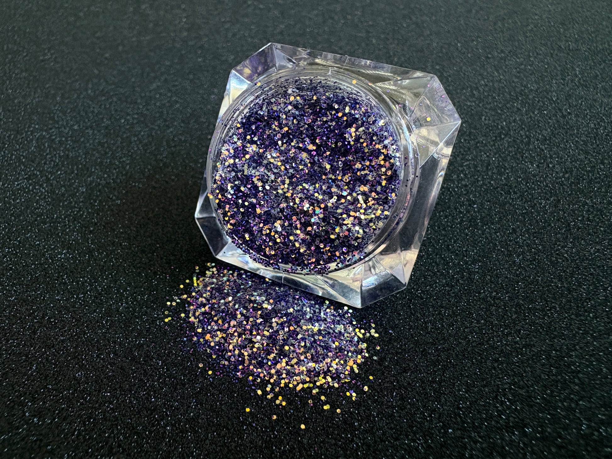 Purple and gold glitter mix spilled from a clear jar onto a black textured background.