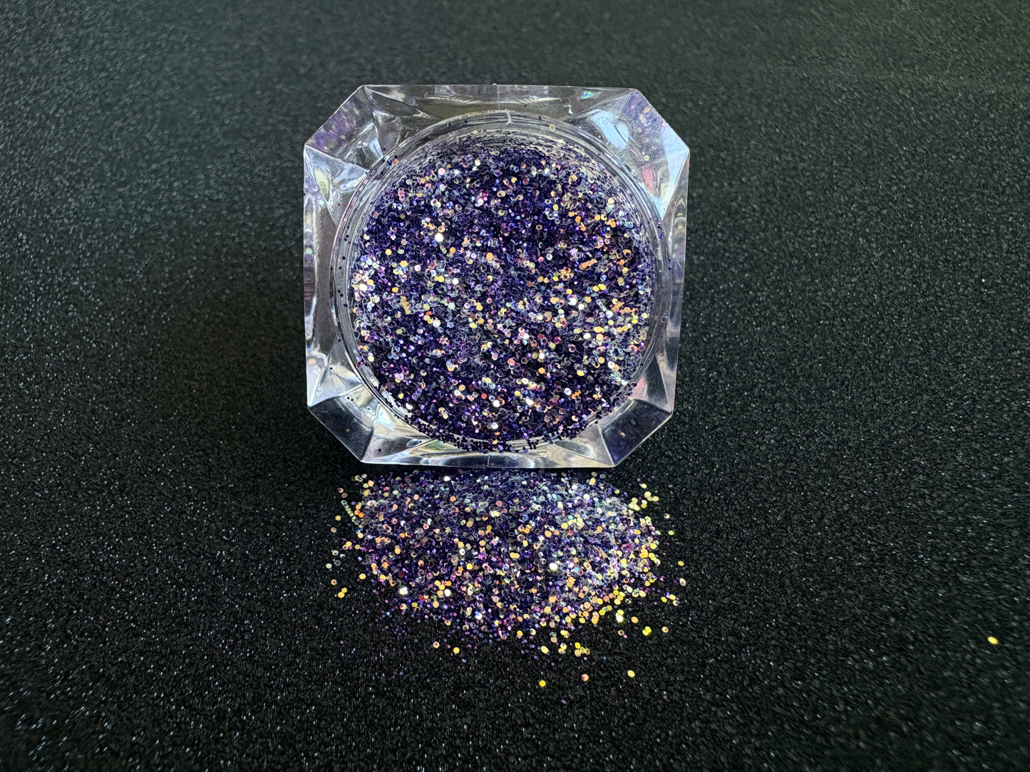 Purple and gold glitter mix spilled from a clear jar onto a black textured background.