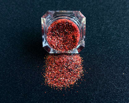 Bright coral and black glitter with holographic and iridescent sparkle spilled from a clear jar onto a black textured background.