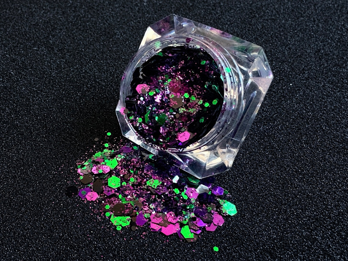 Emerald green, dark purple, and black metallic glitter mix spilled from a clear jar onto a black textured background.