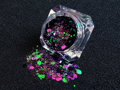 Emerald green, dark purple, and black metallic glitter mix spilled from a clear jar onto a black textured background.