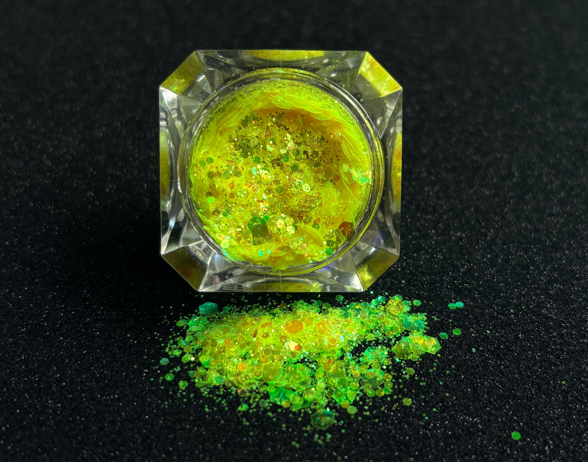 Fluorescent iridescent green super chunky glitter mix with yellow shift spilled from a clear jar onto a black textured background.