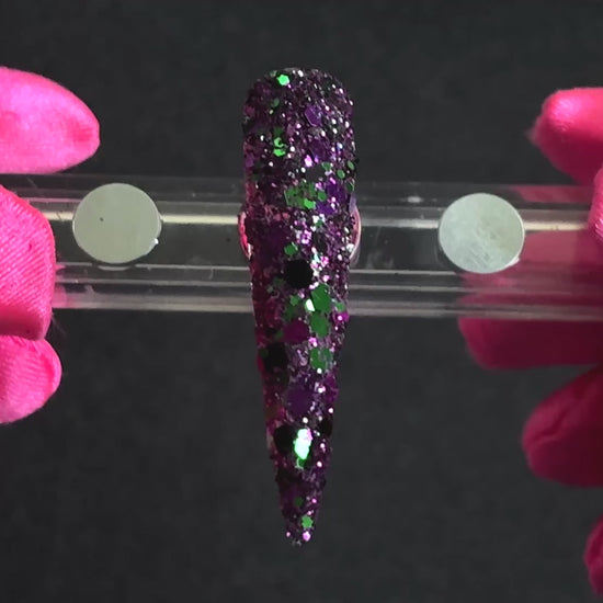 Emerald green, dark purple, and black metallic glitter mix applied on a stiletto nail tip, tilted back and forth to show sparkle.