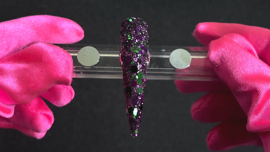Emerald green, dark purple, and black metallic glitter mix applied on a stiletto nail tip, tilted back and forth to show sparkle.