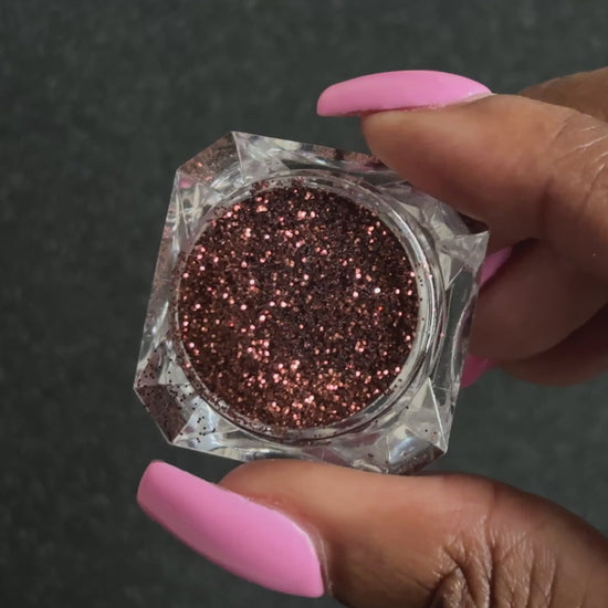 Chocolate brown metallic glitter in a clear jar, tilted back and forth to show sparkle against a black background.