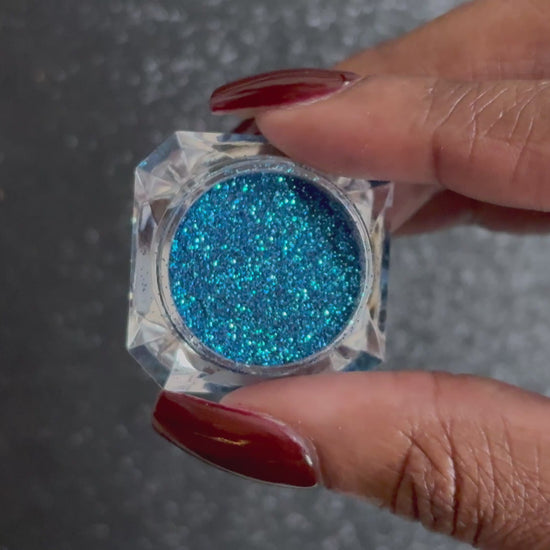 Blue and teal glitter mix in a clear jar, tilted back and forth to show sparkle against a black background.
